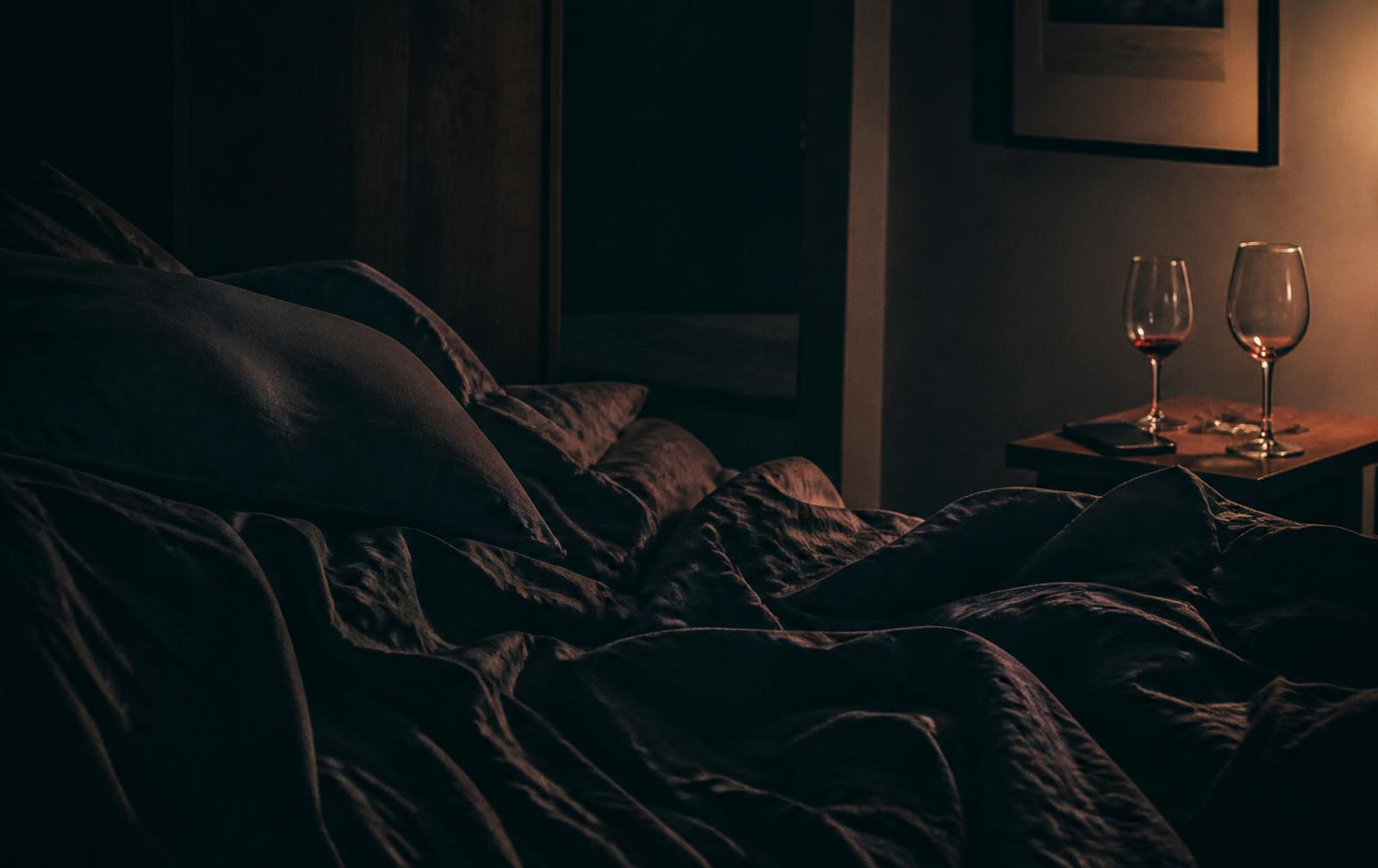 Dim bedroom in low amber light with rumpled dark linens and two glasses on a nightstand